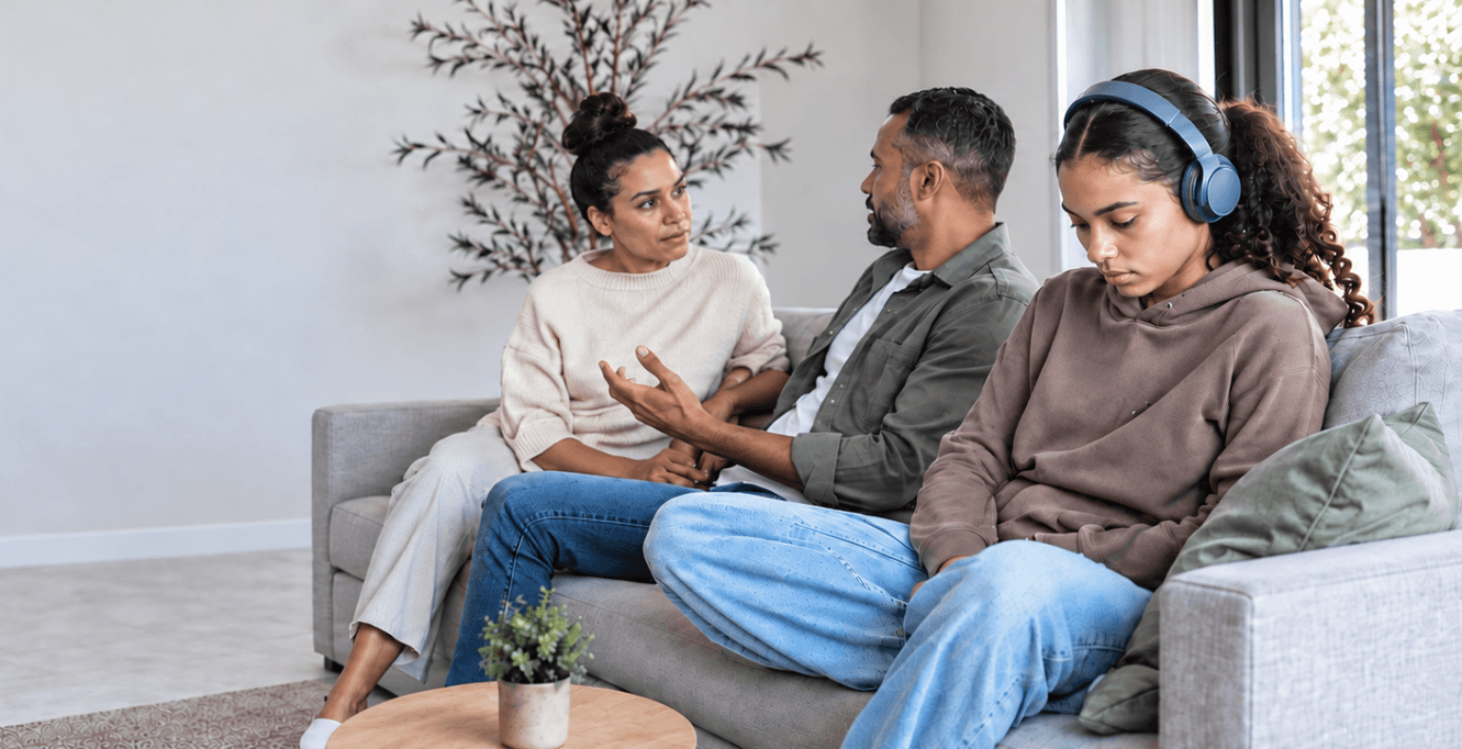 Two adults talk while a teenager wearing headphones sits apart, reflecting the effort families make to understand and support different needs.